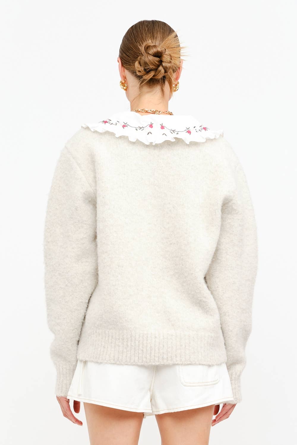 Person wearing a light-colored sweater with a floral collar and white shorts on a plain background